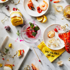 Studio food photography of a post-party scene with empty plates, seafood, cocktails, and scattered garnishes on a marble table.
