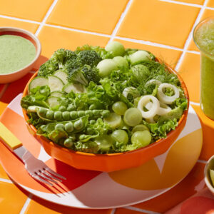 Studio food photography of a monochromatic green salad in an orange bowl, with a green smoothie and dressing on orange tiles.