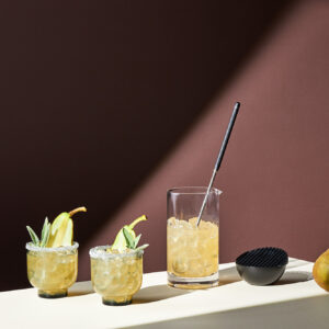 Beverage studio photography with two garnished cocktails, a mixing glass, and a pear on a light surface against a dark background.